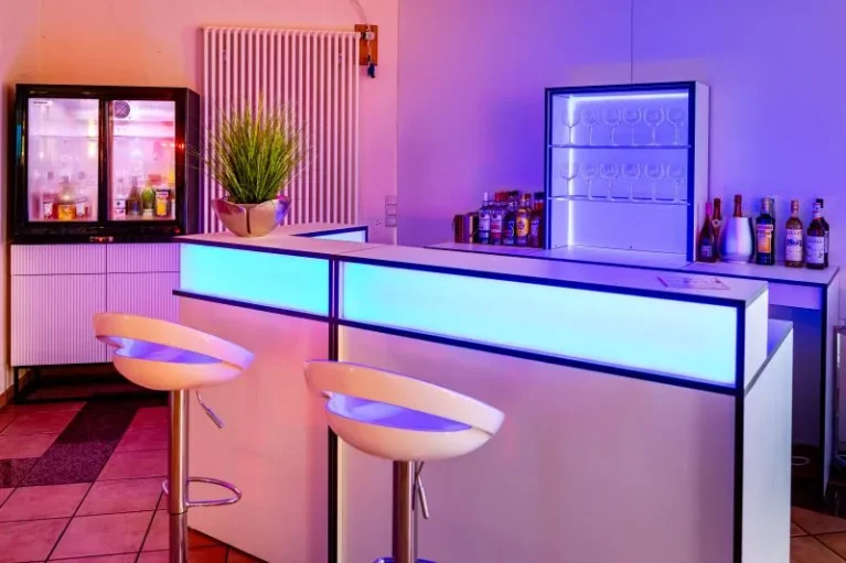 Moderne Hausbar mit LED-Beleuchtung, zwei Barhockern, Getränken und Gläserregal; stilvolles Ambiente für gesellige Abende zu Hause.