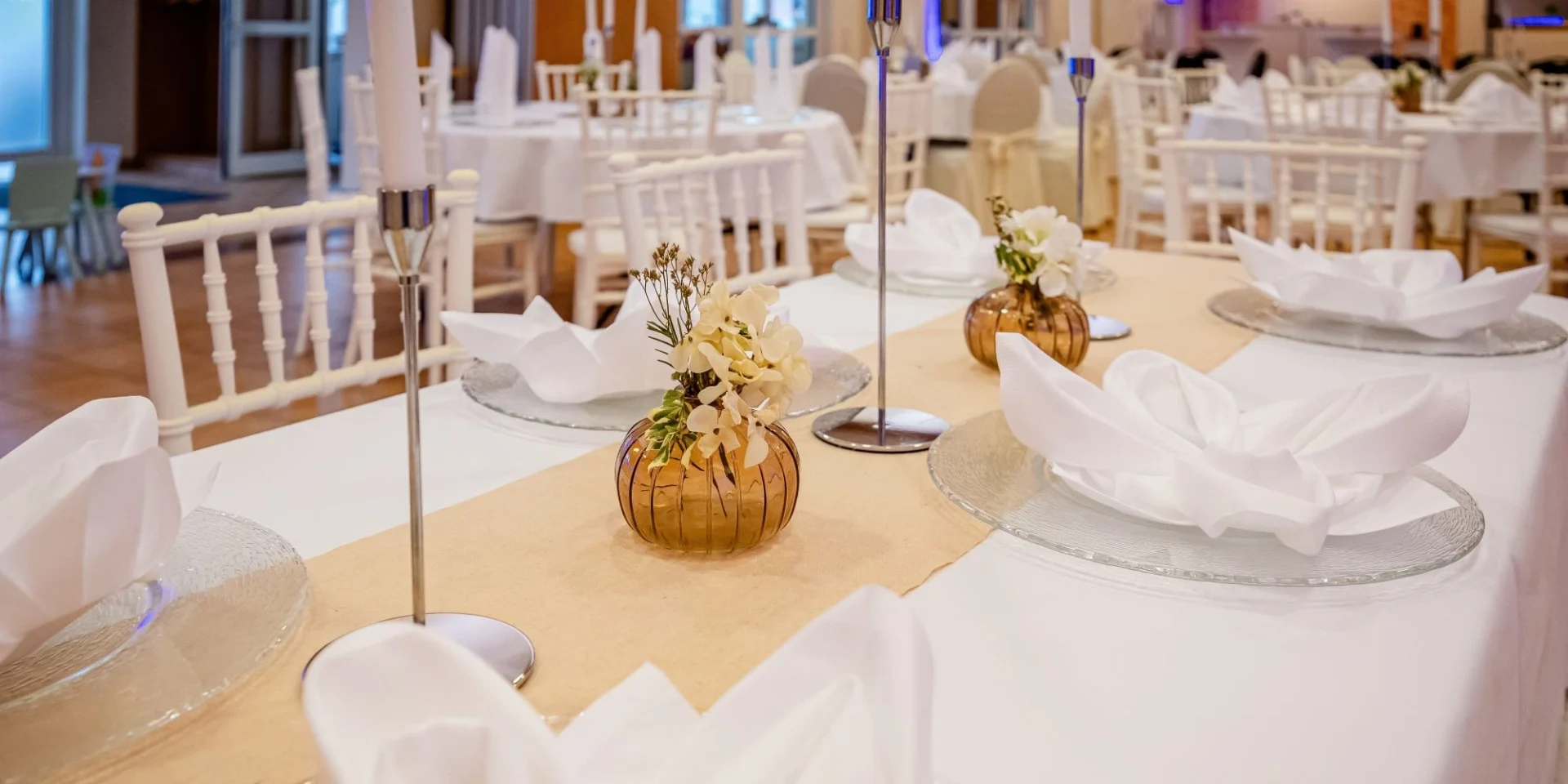 Elegante Hochzeitstischdekoration mit weißen Servietten, Kerzen und Blumen in einem stilvollen Saal.