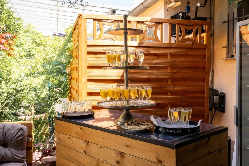 Terrasse mit Holzbar, Sektgläsern auf Etagere und Sonnenschein, umgeben von Grün. Perfekt für einen stilvollen Empfang im Freien.