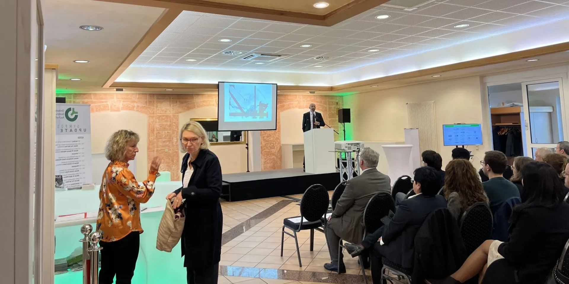 Konferenzraum mit Vortragendem auf Bühne, Publikum sitzt, zwei Frauen im Gespräch im Vordergrund.