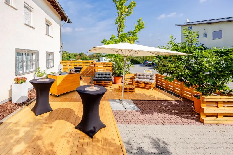 Moderne Terrasse mit Holzdeck, Stehtischen, Sofas aus Paletten und Sonnenschirm, umgeben von grüner Bepflanzung unter blauem Himmel.