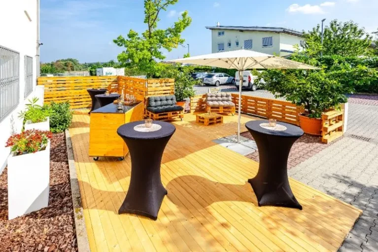 Moderne Outdoor-Terrasse mit Holzboden, Stehtischen, Palettensofas und Sonnenschirm, ideal für entspannte Events.
