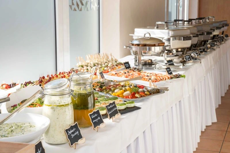 Elegantes Buffet mit vielfältigen Gerichten, Salaten und Dressings auf weißem Tisch in lichtdurchflutetem Raum. Ideal für Veranstaltungen.