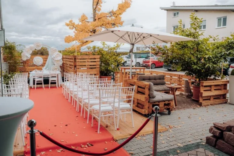 Außentrauungsbereich mit roten Teppich, weißen Stühlen, Sofa aus Paletten und herbstlicher Dekoration für Hochzeit.