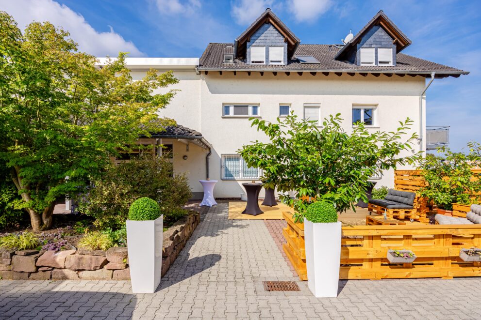 Moderne Terrasse mit Lounge-Möbeln aus Paletten, dekoriert mit Pflanzen und Stehtischen vor zweigeschossigem Wohnhaus.