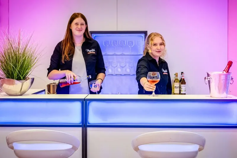 Zwei Barkeeperinnen in einer beleuchteten Bar servieren Drinks, umgeben von farbigem Licht und dekorativen Elementen.