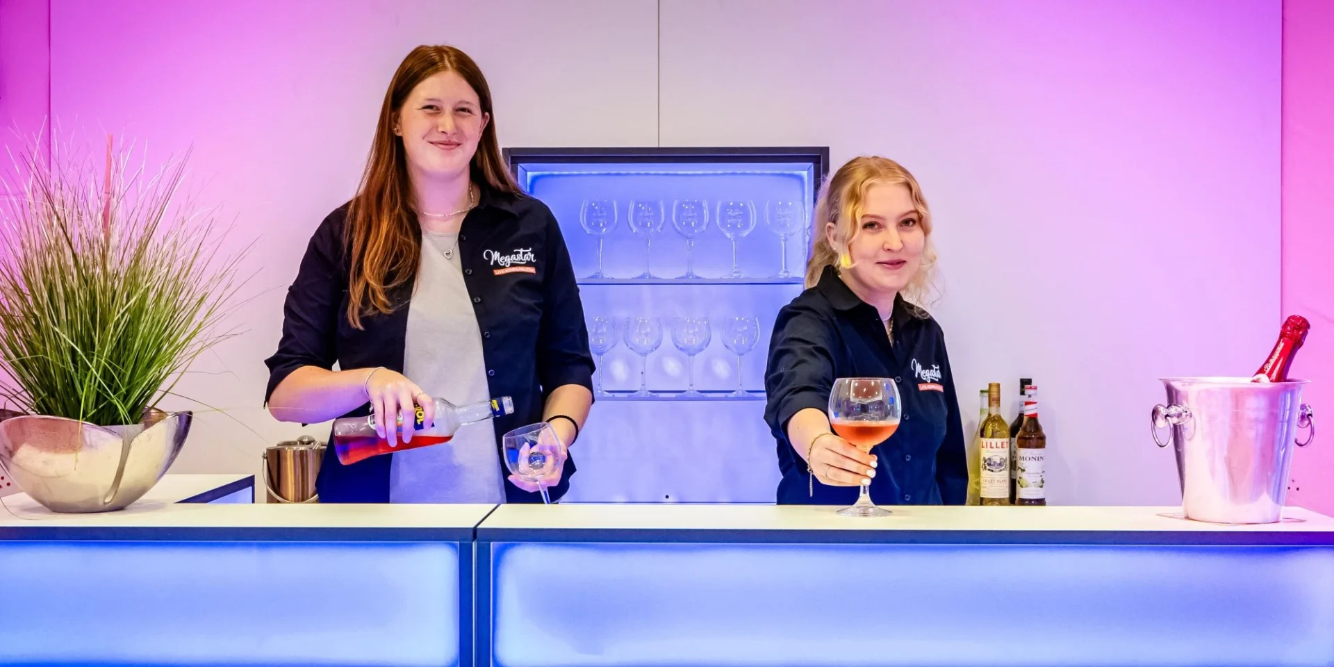 Zwei Frauen hinter einer Bar schenken Getränke ein, umgeben von Flaschen und Beleuchtung in Pink und Blau.