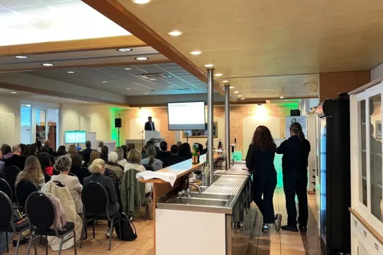 Vortrag mit Publikum in einem Seminarraum, Redner am Podium, Getränkeausgabe im Vordergrund.