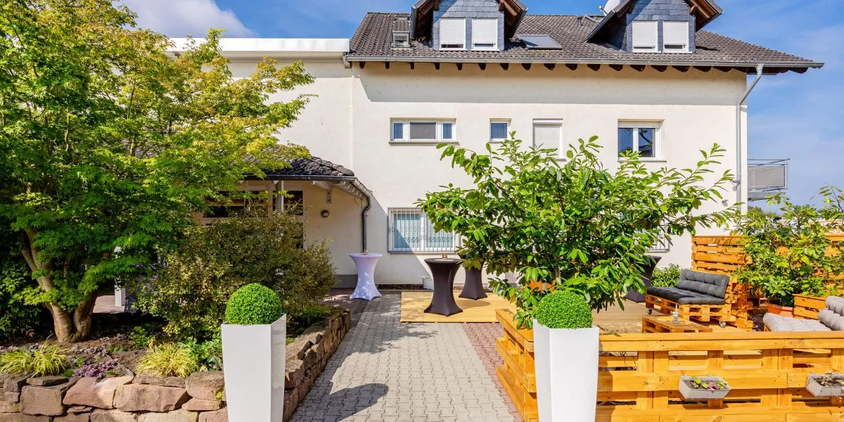 Moderner Außenbereich mit Holzterrasse, weißen Pflanzgefäßen und Sitzbereich vor einem weißen Haus bei sonnigem Wetter.